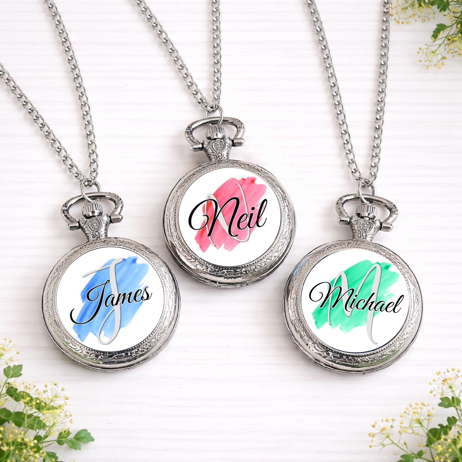 Three silver personalised pocket watches featuring custom names over coloured brushstroke backgrounds in blue, pink and green.